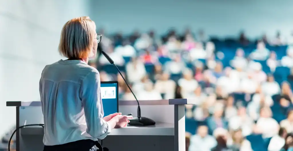 Female speaker giving a talk on corporate business conference. Unrecognizable people in audience at conference hall. Business and Entrepreneurship event