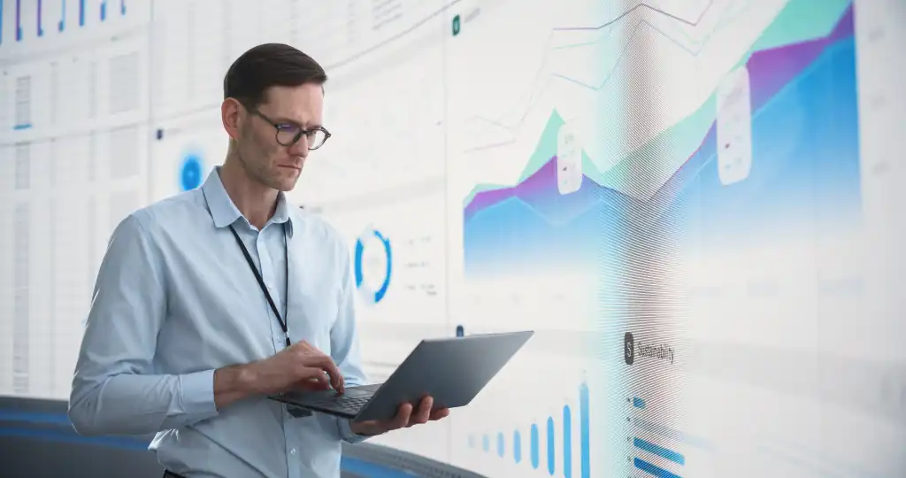 Caucasian Male Business Analyst With Laptop Computer Standing Next To Big Digital Screen With Graphs And Charts. Successful Man Assessing Risks And Developing Strategies For Company Growth, Expansion.