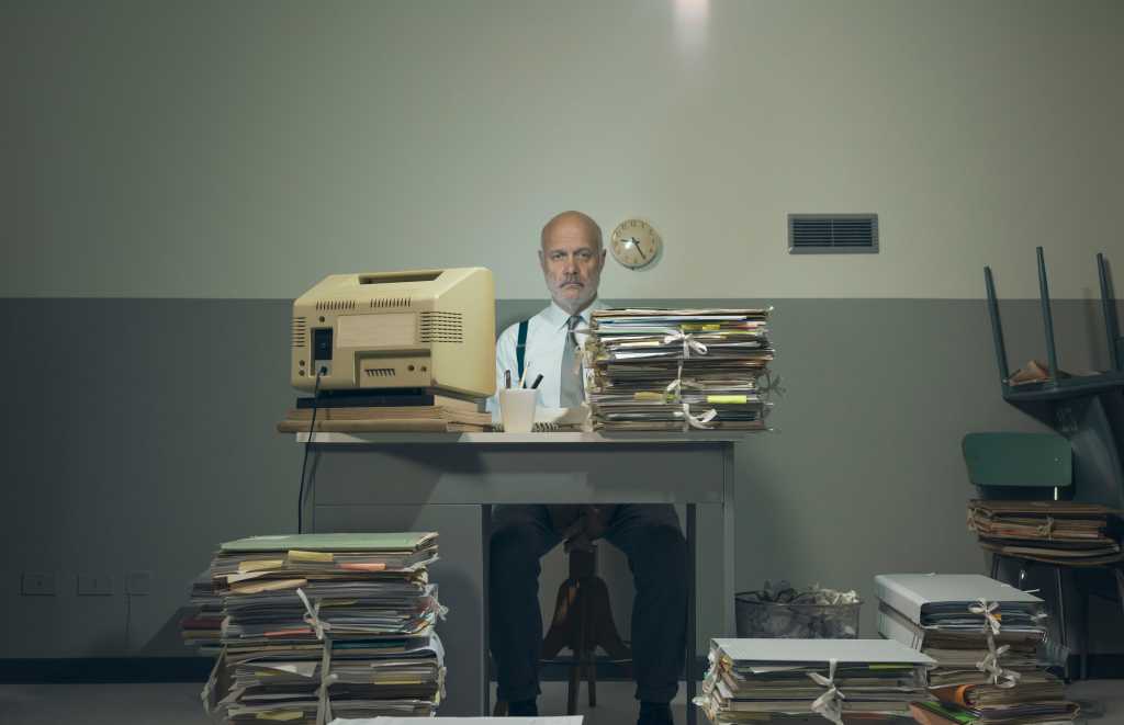 Frustrated vintage style businessman working in a rundown old office space, he is overloaded with papework